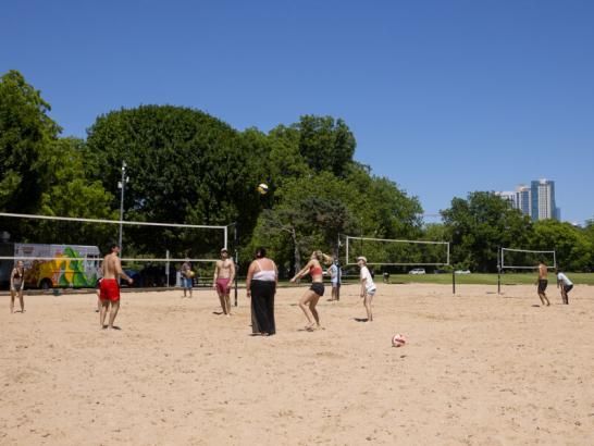 People play sand volleyball in Zilker Park. A part of the downtown Austin skyline is visible behind trees in the background.