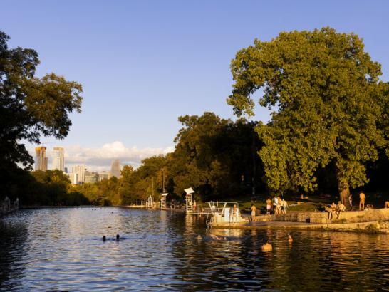 People swim and sunbathe at sunset at Barton Springs. A view of the downtown Austin skyline is visible behind trees in the background.