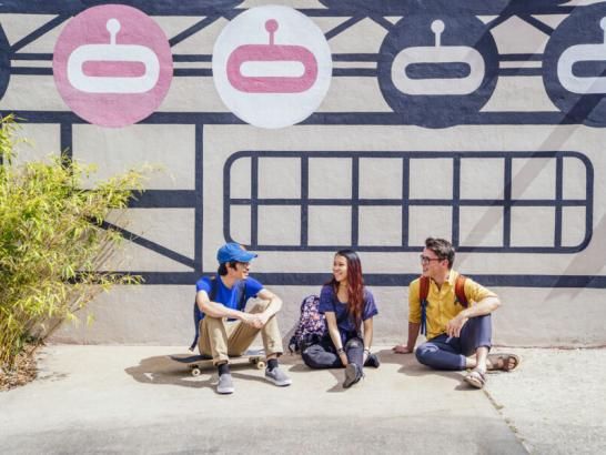Three students sit on a sidewalk on South Congress Avenue in front of a black, white and pink mural.