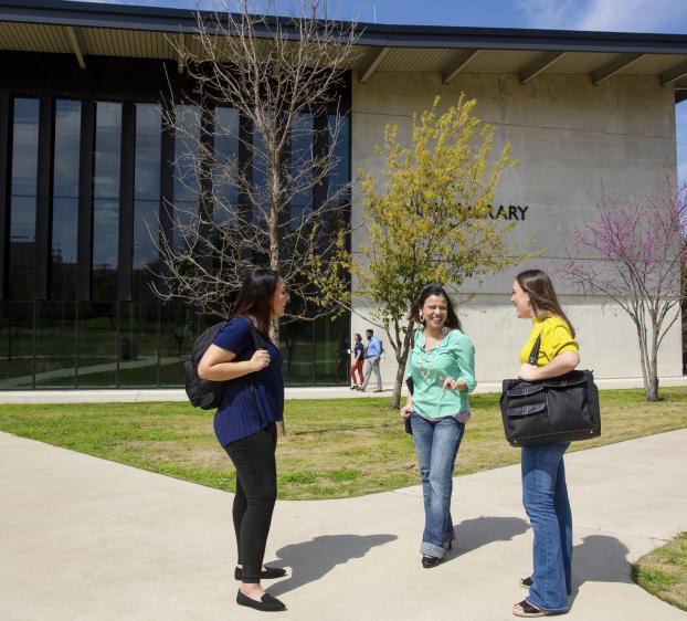 Three graduate students stand together on a pathway outside of Munday Library and chat.