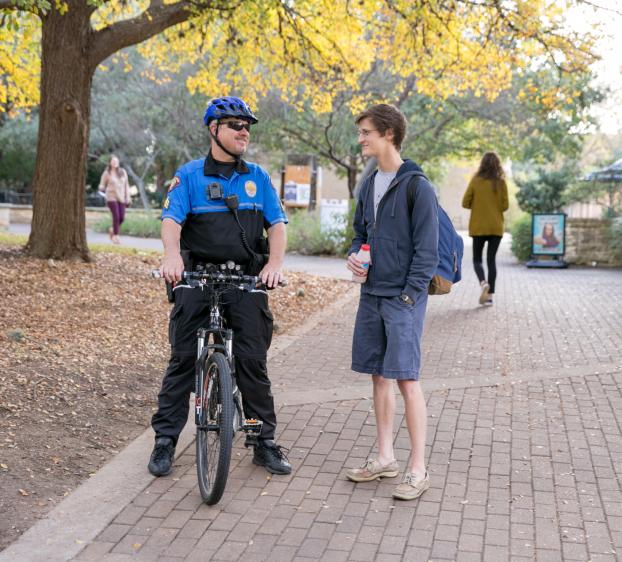 A university police officer sits on their bicycle and talks to a student as other students walk behind them.