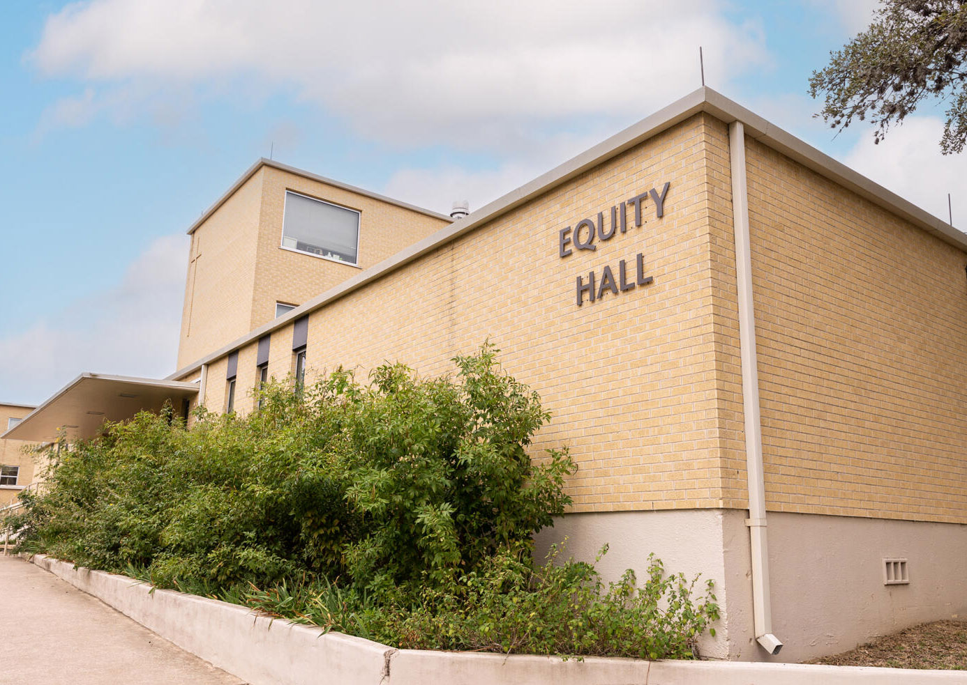 The image shows the exterior of a building named "Equity Hall." The structure is made of light yellow brick and has a modern, rectangular design. The name "Equity Hall" is prominently displayed in large, bold letters on the side of the building. In front of the building, there are well-maintained shrubs and a paved walkway. The sky above is partly cloudy, giving the scene a bright and calm atmosphere.