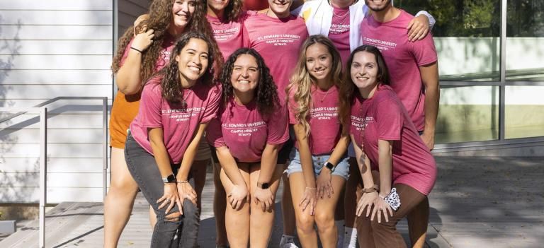 Students wearing Campus Ministry retreat shirts stand in two lines for a group photo outside of the Campus Ministry building.