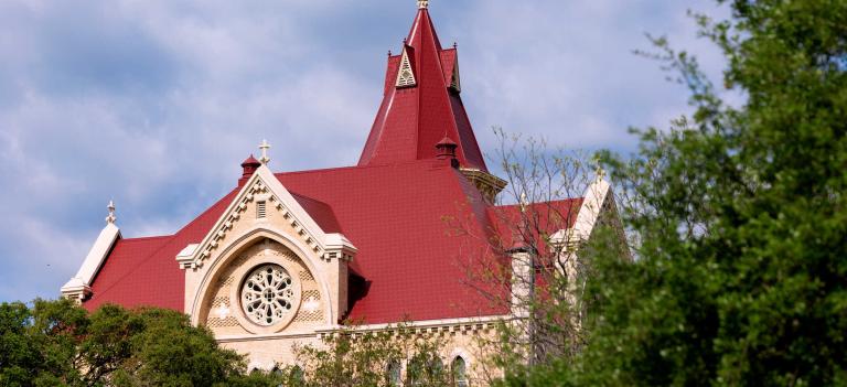 Main Building's steeple and roof from the side, framed by tree branches in the foreground.