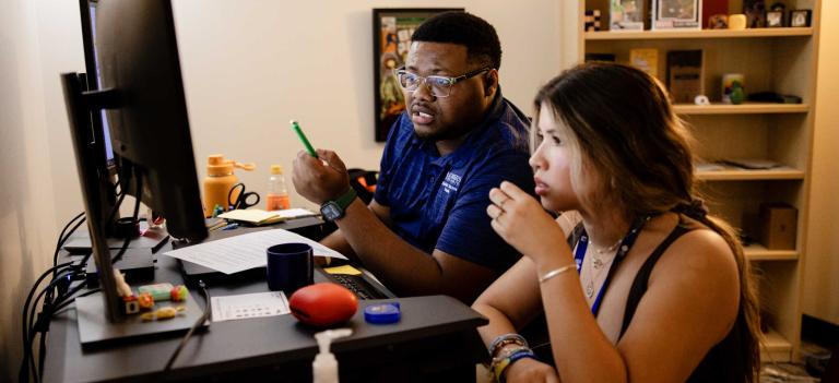 A success coach and a student meet in the success coach's office. They sit at the success coaches desk and look at the computer screen the success coach is pointing at with a pen.