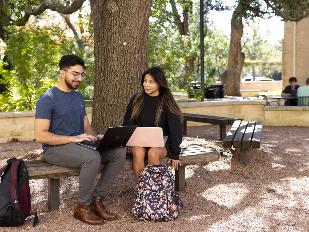 Two students sit under a tree on a bench on a patio with their backpacks on the ground next to them and work on their computers.