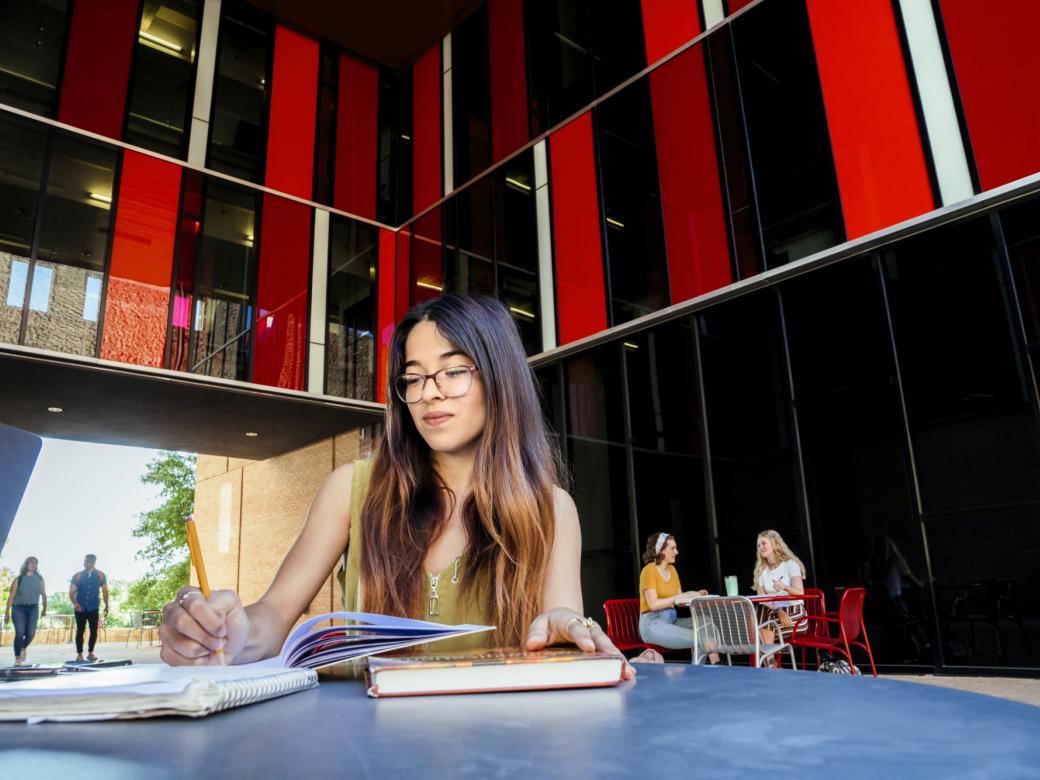 A student sits at a table and writes in a notebook. Iconic red and black architecture of The Village frame her in the background.