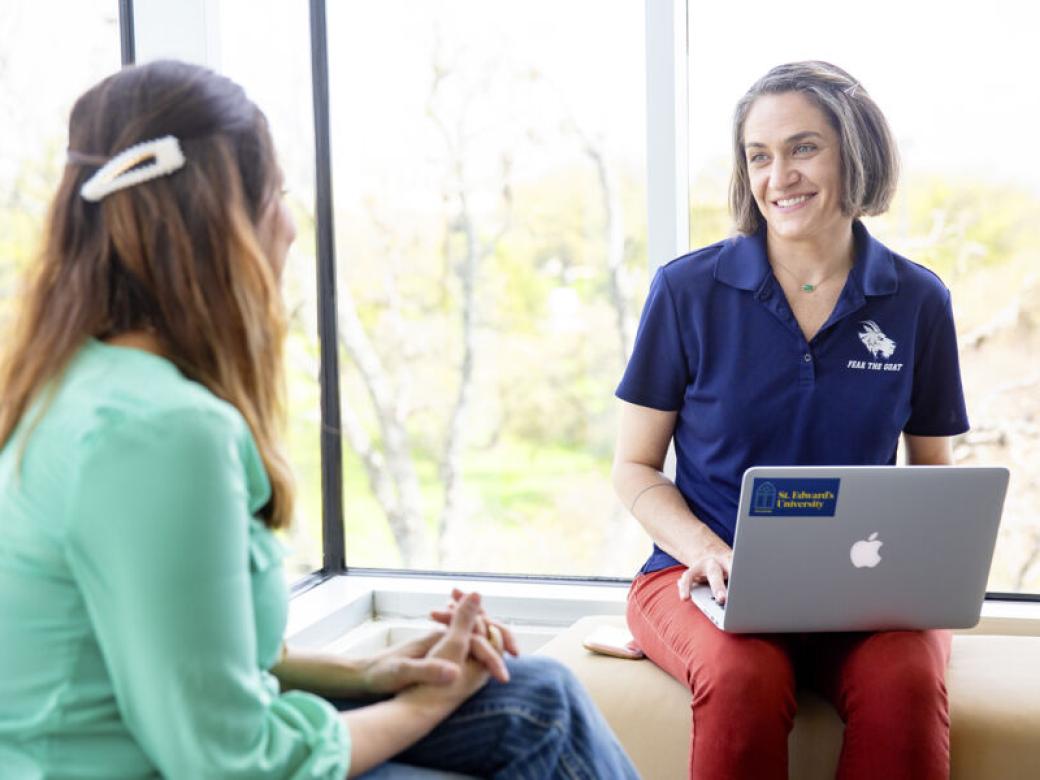 Graduate students sit on benches by a window and talk. One student is wearing a St. Edward's polo and has a laptop in their lap.