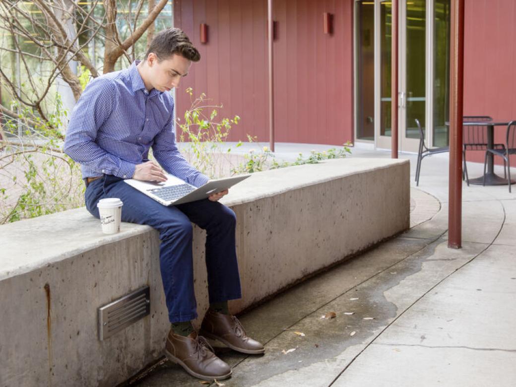 A graduate student sits on a concrete bench looking at their laptop with a cup of coffee next to them on the John Brooks Williams Natural Sciences Center - South patio.