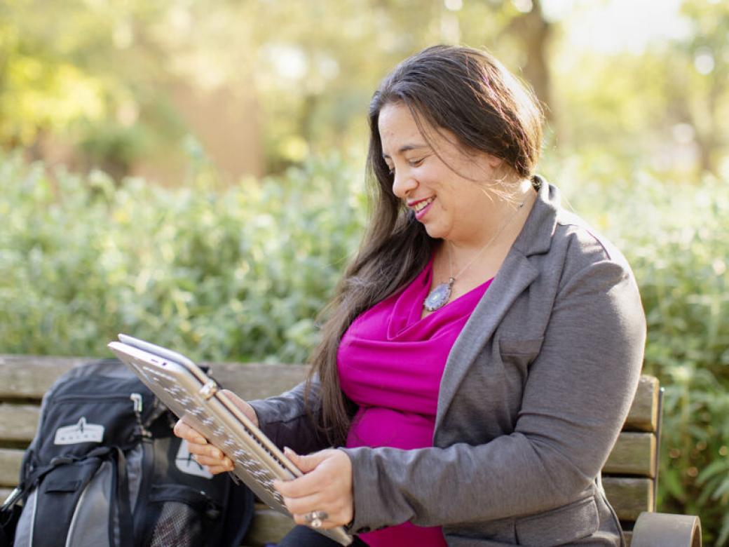 A graduate student sits on a bench with their backpack next to them holding a laptop in a tablet orientation.