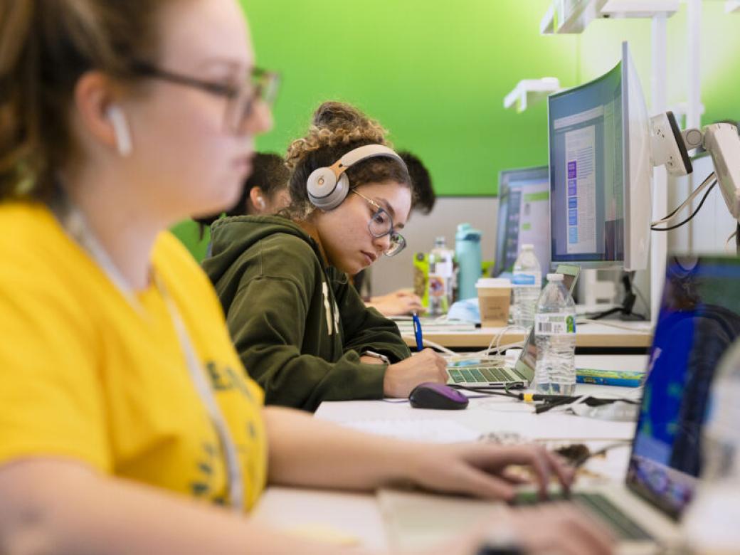 A student wearing headphones takes notes while working at a computer and is framed by other other students working at computers.