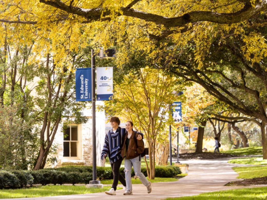 Two students walk arm-in-arm on a tree-lined path showing fall foliage, with St. Edward's banners on lightpoles behind them.