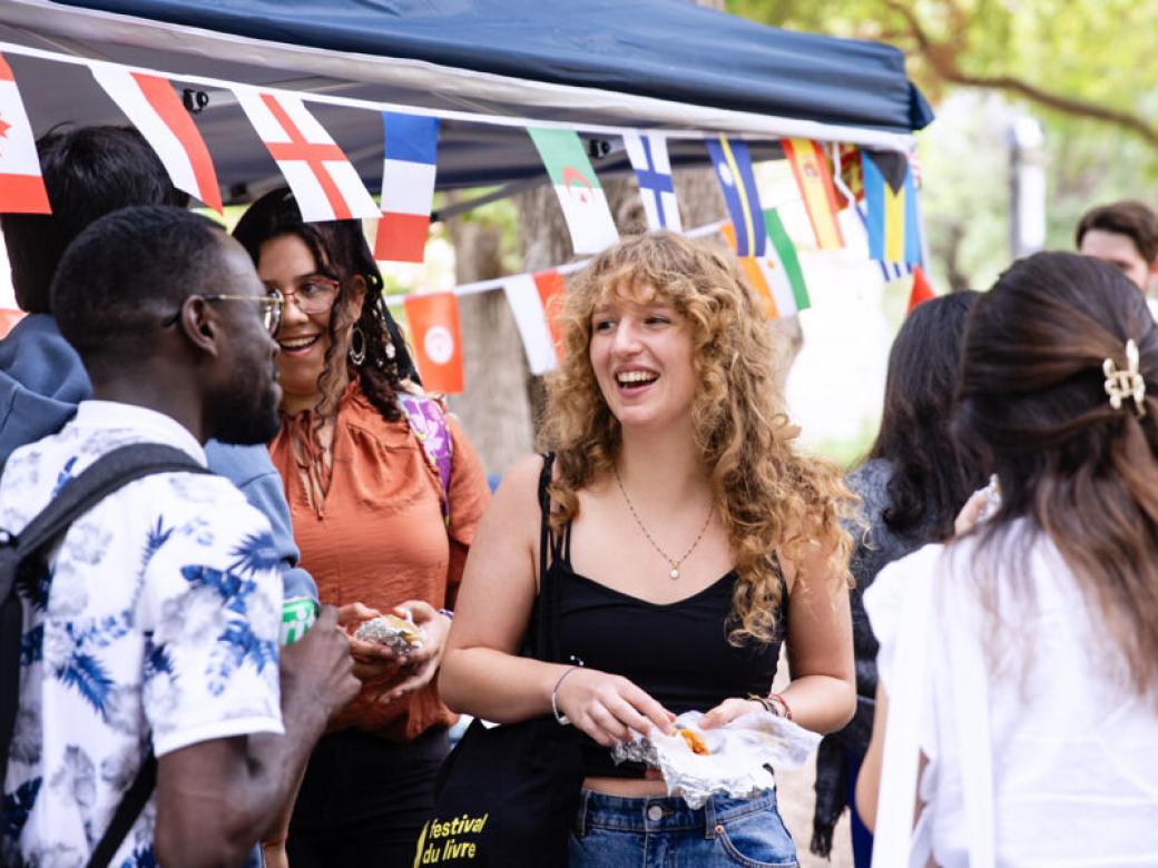 International students mingle and eat snacks by a tent displaying different countries' flags during an international student event.