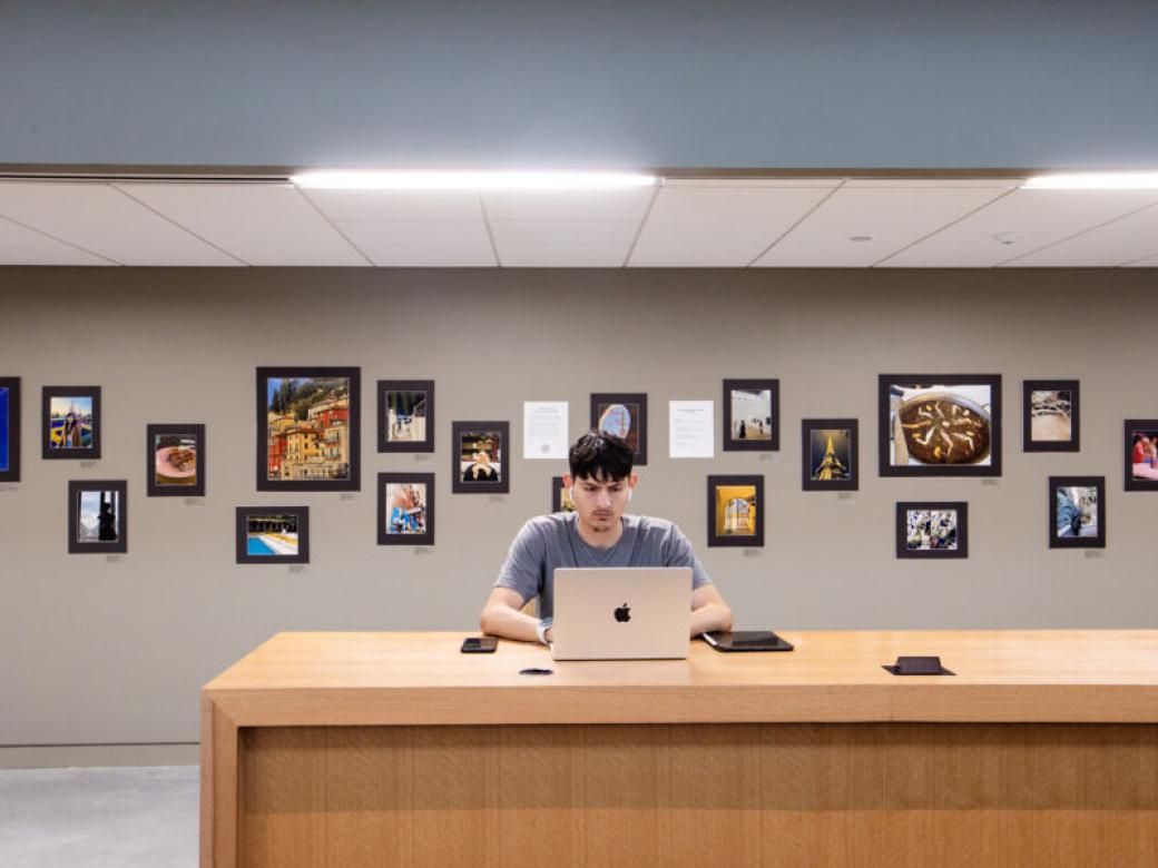 A student sits at a long desk and types on their laptop. A gallery of photos on black mattes are on the wall behind them.