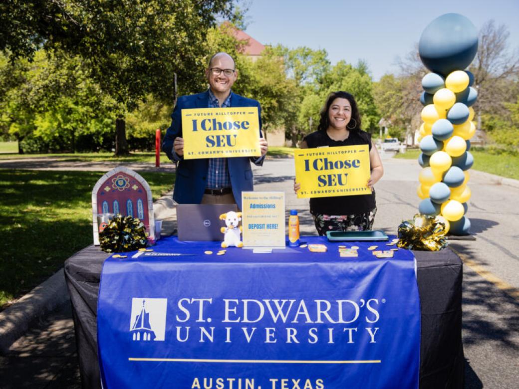 Two admission counselors stand at a table with A St. Edward's University banner on it and hold I Chose SEU signs.