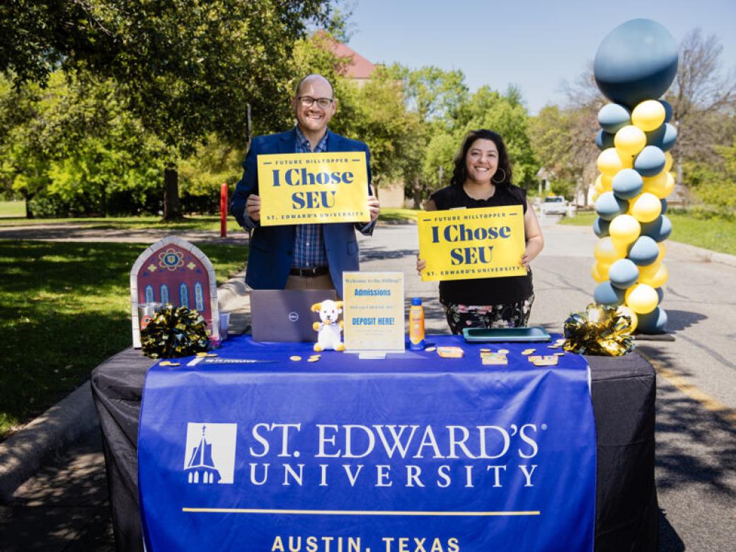 Two admission counselors stand at a table with A St. Edward's University banner on it and hold I Chose SEU signs.