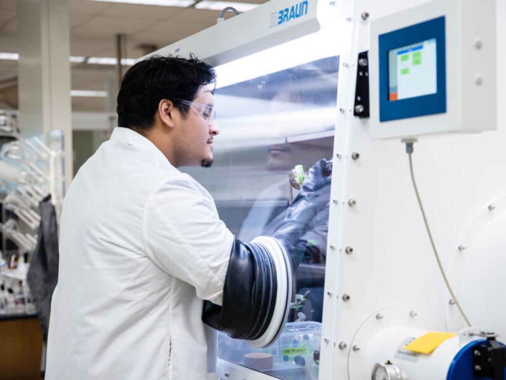 A student wearing a lab coat and protective glasses looks at bottles of chemicals using specialized equipment.