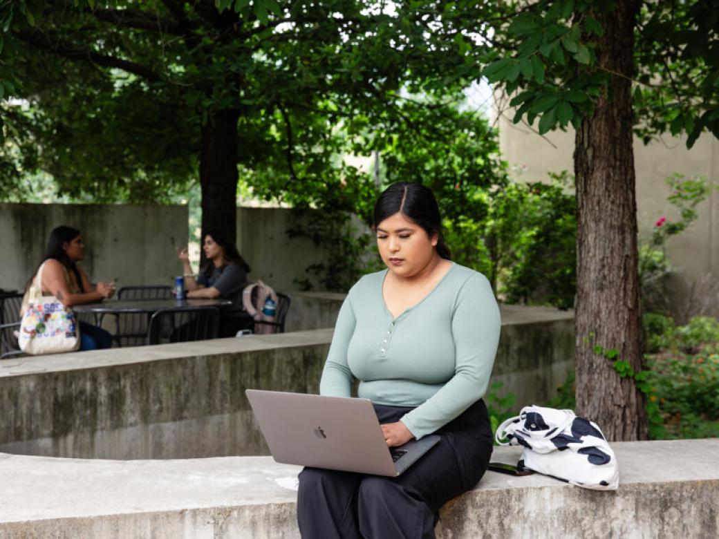 A student sits outside with a laptop on their lap and types. Trees and two other students sitting at a patio table are in the background.