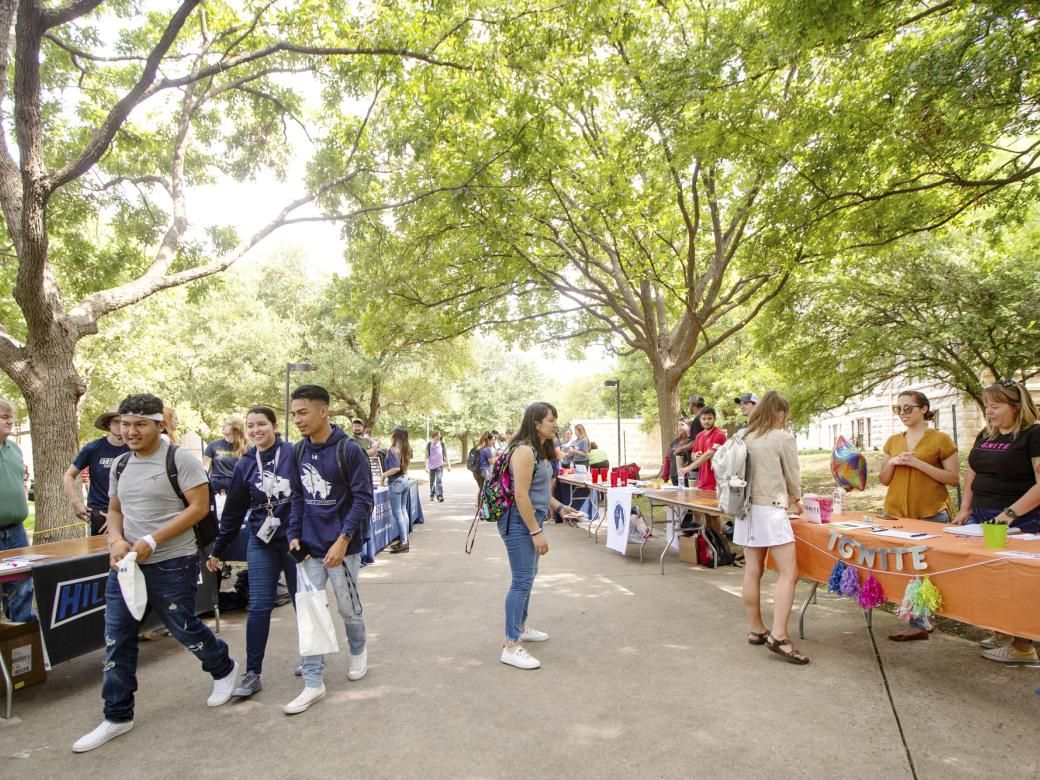 Students walk down a path with a tree canopy overhead. The path is lined by tables as students stop to get information from other students, faculty and staff.