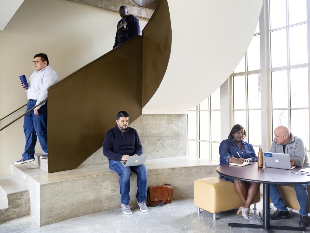 A student walks down the stairs. Another student sits on a bench. Two students sit at a table. All inside The Bill Munday School of Business.