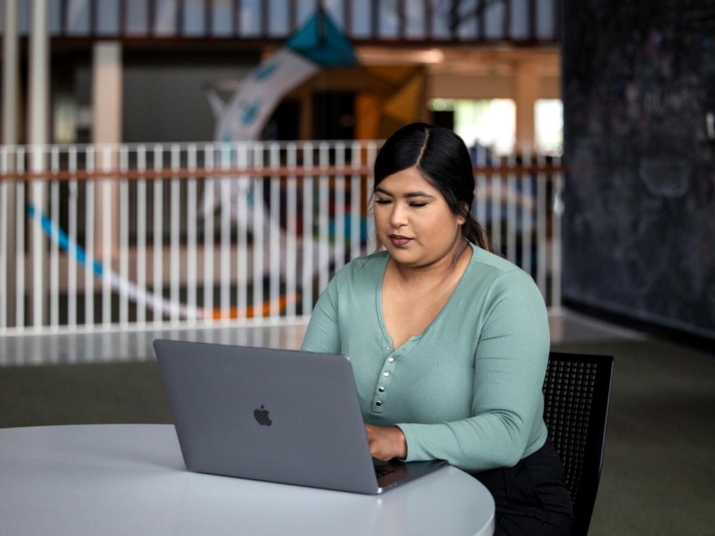 A student sits at a table in the Munday Library and types on their laptop.