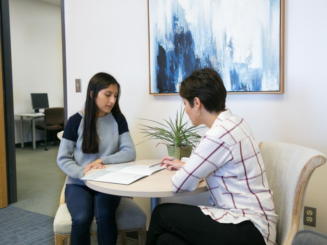 A student and a financial aid counselor sit at a table in an office and look over financial information.