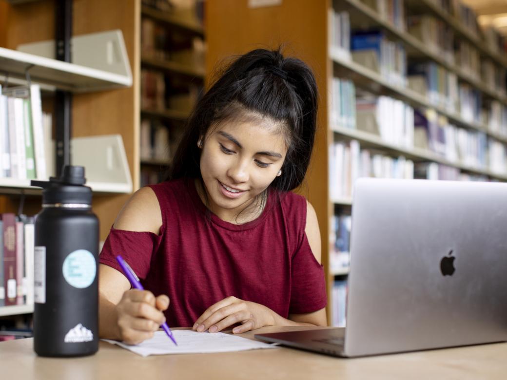 A student sits at a table with a water bottle and a laptop and takes notes on a piece of paper. Bookcases are in the background.