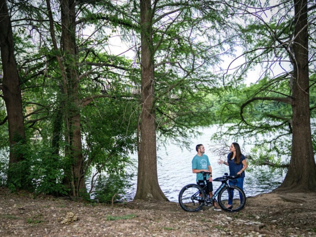 A student and a professor stand by Lady Bird Lake with a bike and are surrounded by trees.