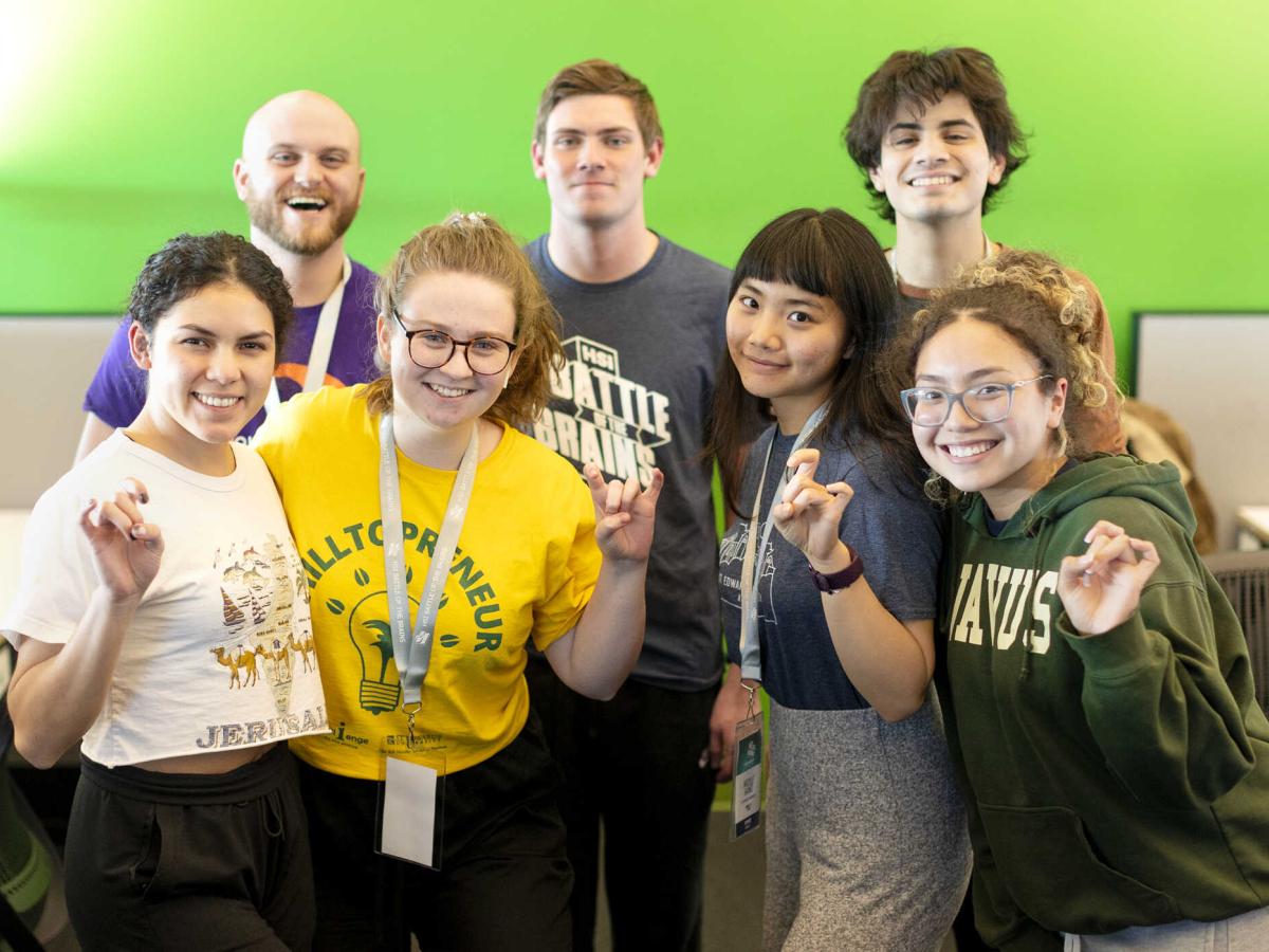 Seven students stand in front of a green wall and give toppers up hand signs.