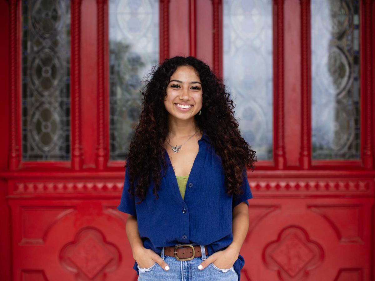 A young woman with curly hair stands smiling in front of an ornate red door with decorative glass panels. She is wearing a blue button-up shirt over a green top, paired with denim jeans. Her hands are in her pockets, and she is accessorized with a butterfly necklace. The vibrant red of the door contrasts with her outfit, creating a striking background for the portrait.