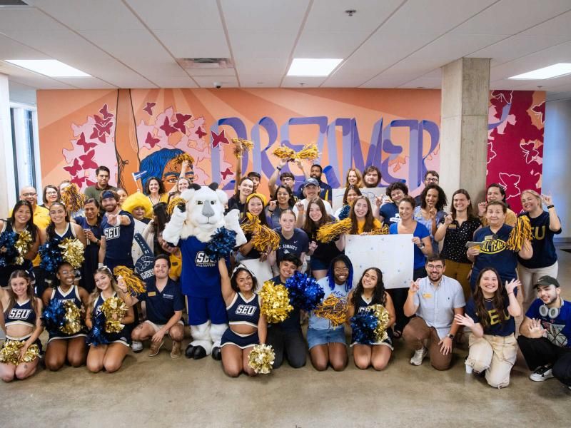 The image depicts a large group of people, including cheerleaders, faculty, and a mascot, posing enthusiastically in front of a colorful mural with the word "DREAMER" prominently displayed. The group is dressed in blue and gold, with many holding pom-poms, and includes the SEU (St. Edward's University) mascot. The atmosphere is festive and celebratory, suggesting a school or community event.