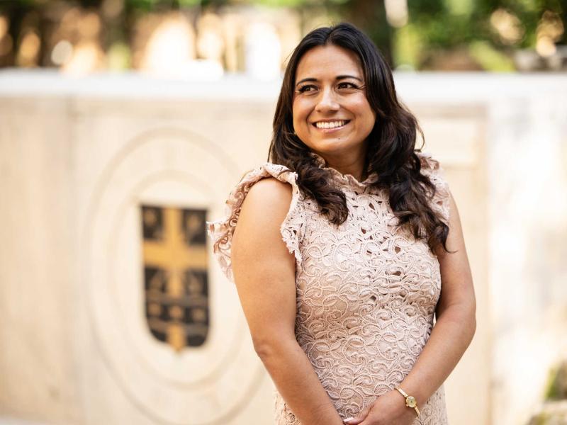 The image shows a woman with long dark hair wearing a light pink lace dress, smiling warmly while standing outdoors. In the background, there is a blurred emblem or crest on a stone structure. The setting appears to be a formal or professional environment, possibly a campus or an institution. The woman looks composed and confident.