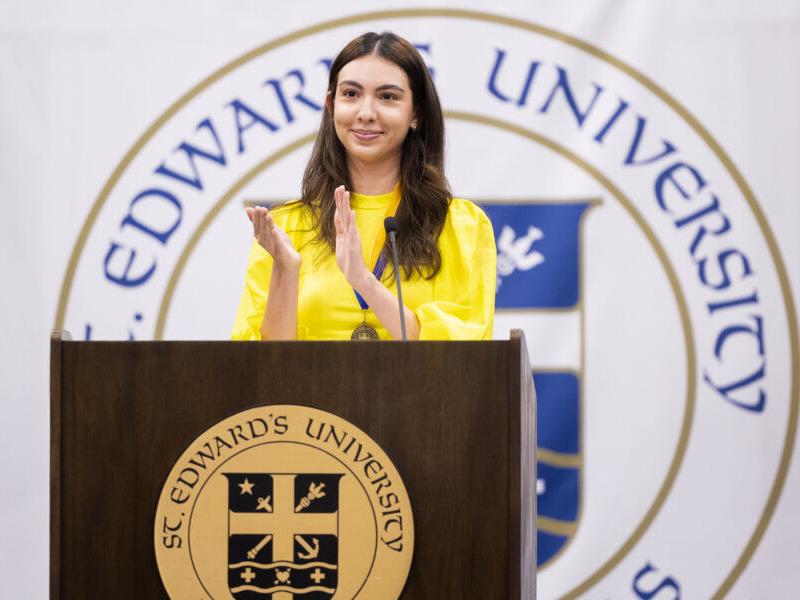 The image shows a woman standing at a podium with the logo of "St. Edward's University" in the background. She is wearing a bright yellow top and is clapping with a smile on her face. The podium has the university's emblem on the front. She appears to be addressing or acknowledging an audience, possibly during a formal event or ceremony at the university.