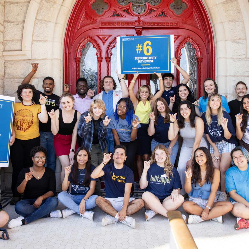 A group of St. Edward's University students pose in front of the red doors with the university president holding up signs celebrating the #6 best university in the west ranking