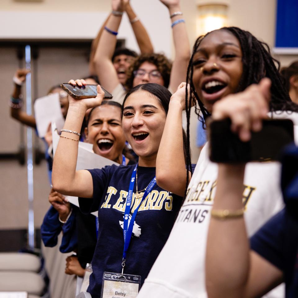 A row of students stand, raise their hands and cheer and wear St. Edward's gear.