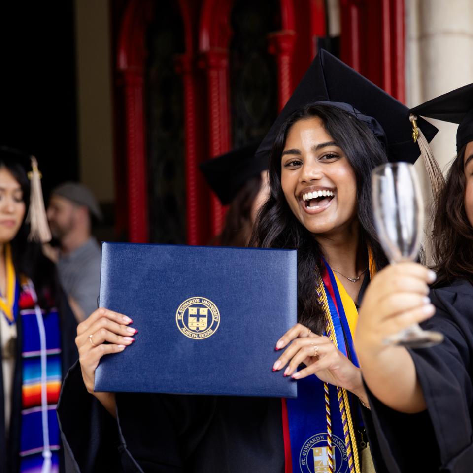 A graduate wearing their cap and gown walks through the red doors of Main Building with fellow graduates, holding their degree and smiling at the camera.