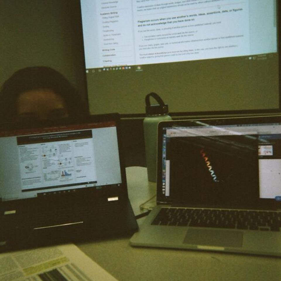 Two laptops on a table with a projector screen behind them.