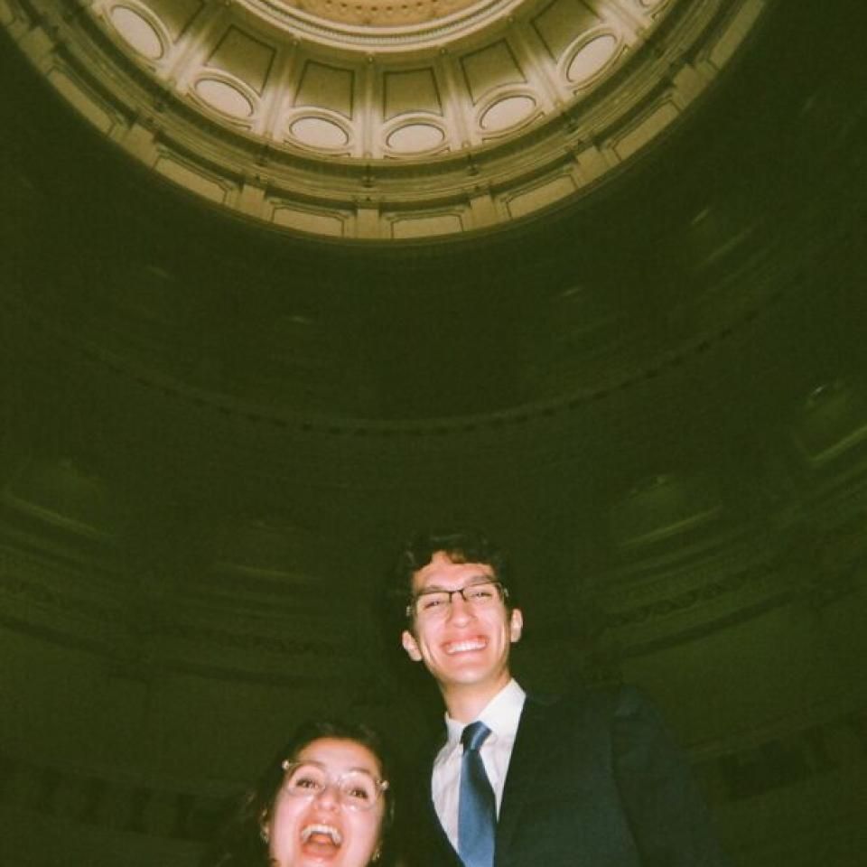 Two people stand for a photo, with the view of the Texas State Capitol rotunda above them.