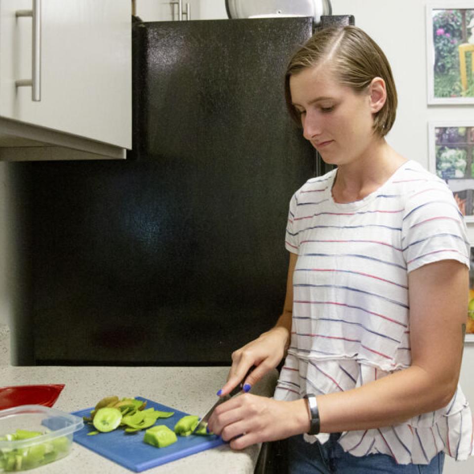 This image shows a woman with short brown hair, wearing a white blouse with red and blue stripes. She is standing in a kitchen, slicing green vegetables on a blue cutting board placed on a countertop. A container with chopped vegetables and a red lid is nearby. Behind her is a black refrigerator, and the wall has a series of colorful framed photographs depicting various fruits and vegetables. The setting appears to be a well-lit, clean, and organized kitchen.