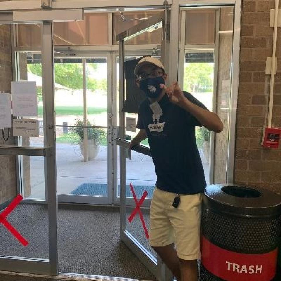 A person wearing a mask holds a door open and makes a toppers up sign.