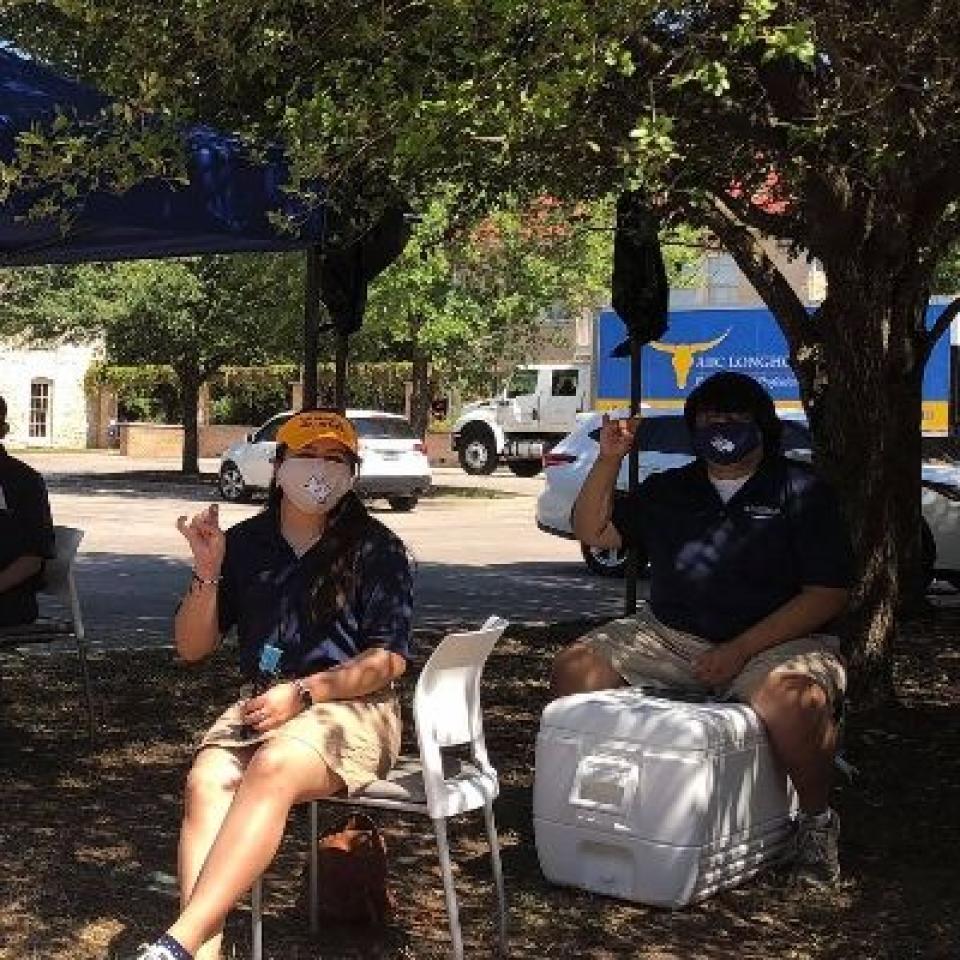 Two people sit outside wearing face masks and giving a toppers up hand sign.