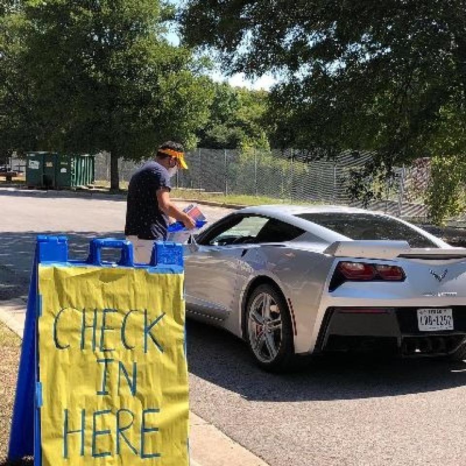 A volunteer wearing a face mask stands by a check in here sign and assists a person in a car.