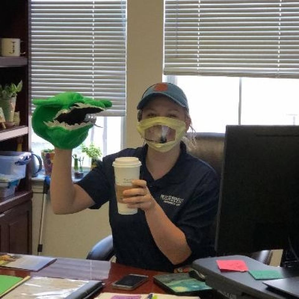 A staff member wears a face mask and holds up a hand puppet and a cup of coffee.