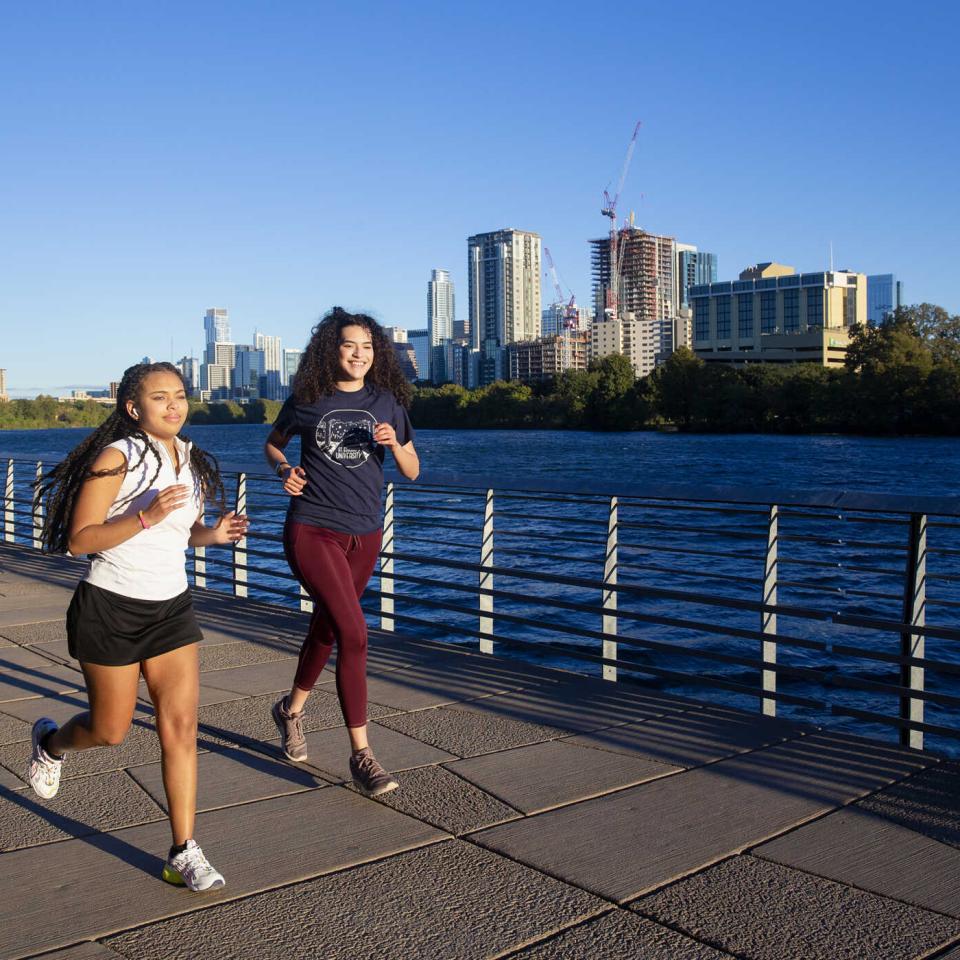 Two stdudents wearing athletic clothing run on the boardwalk of Lady Bird Lake with the lake and the downtown skyline in view at sunrise.