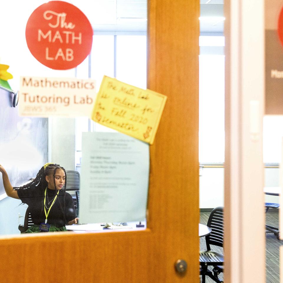 Taylor Huey is seen through the glass of a door as she points to a math problem on a chalkboard.