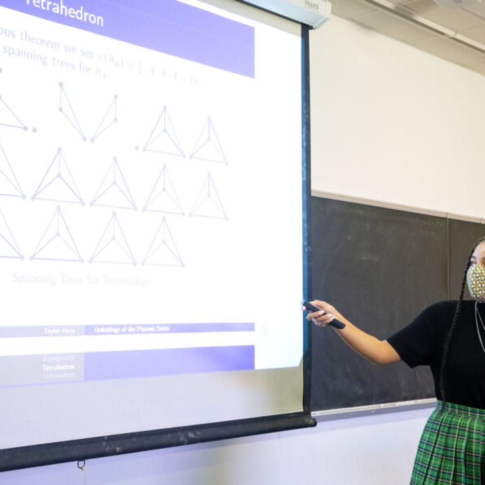 Taylor Huey wears a face mask and stands in front of a projector showing a Powerpoint slide with data as she presents her work.