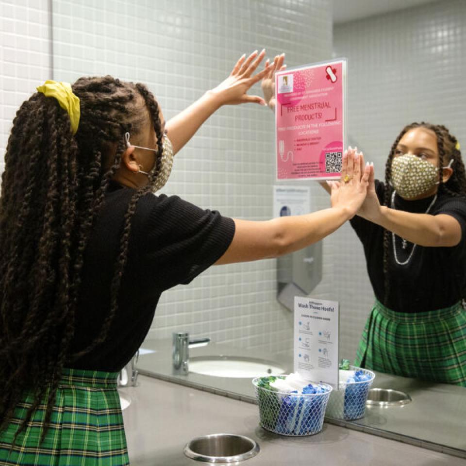 Taylor Huey wears a face mask and puts up a pink sign on a bathroom mirror. She is reflected in the mirror.