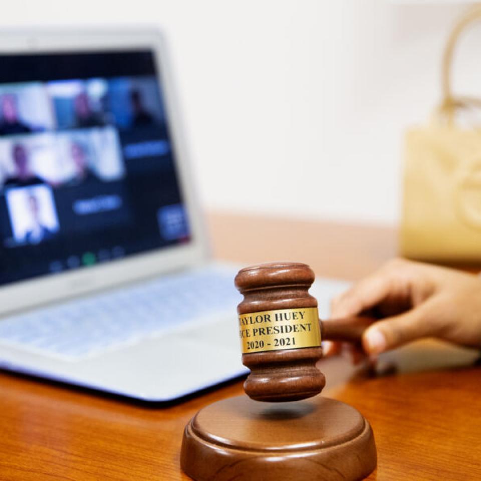 A detail of Taylor Huey's gavel as she holds it and a view of her laptop with SGA members on Zoom.