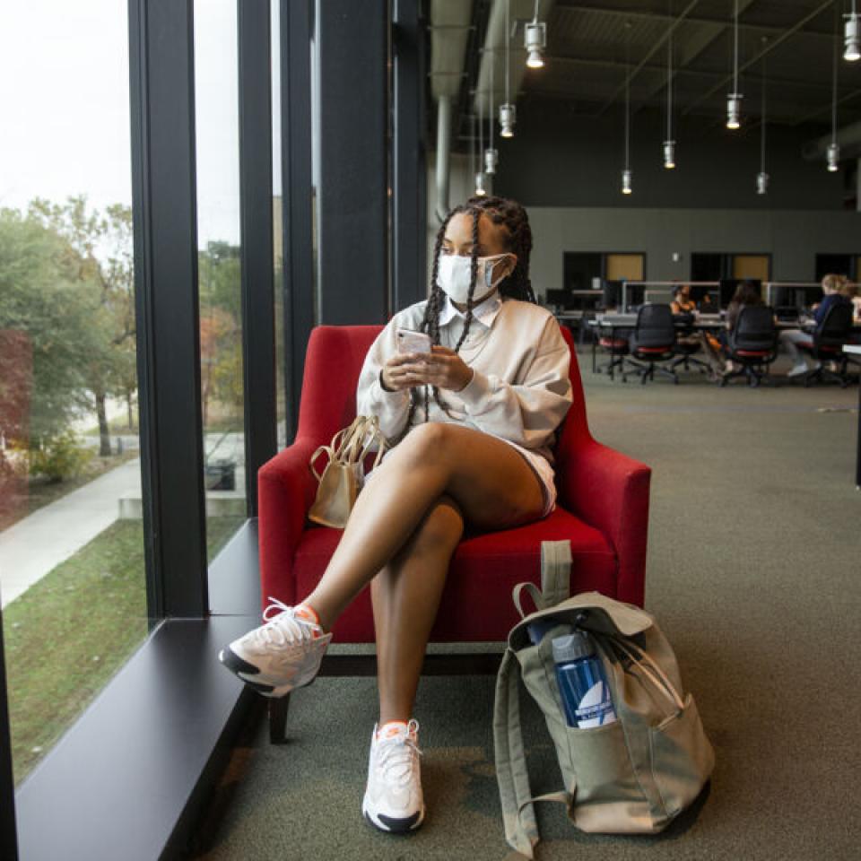 Taylor Huey wears a face mask and sits in a red chair in the Munday Library next to a window. She scrolls on her phone and her backpack is beside her on the ground.