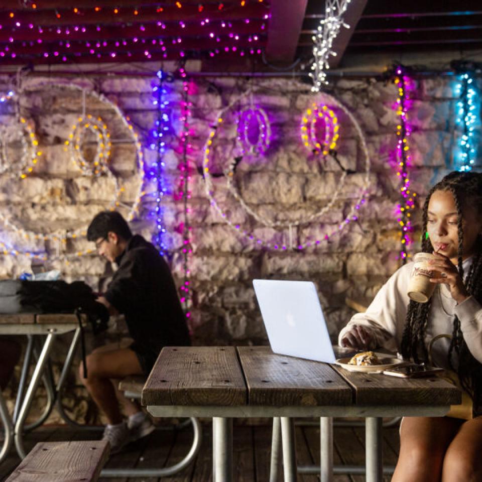 Taylor Huey sits at a picnic table on a covered patio at Mozart's coffee and sips a coffee as she scrolls on her laptop. Multicolor string lights, some in the shape of smiley faces, are on the stone wall behind her.